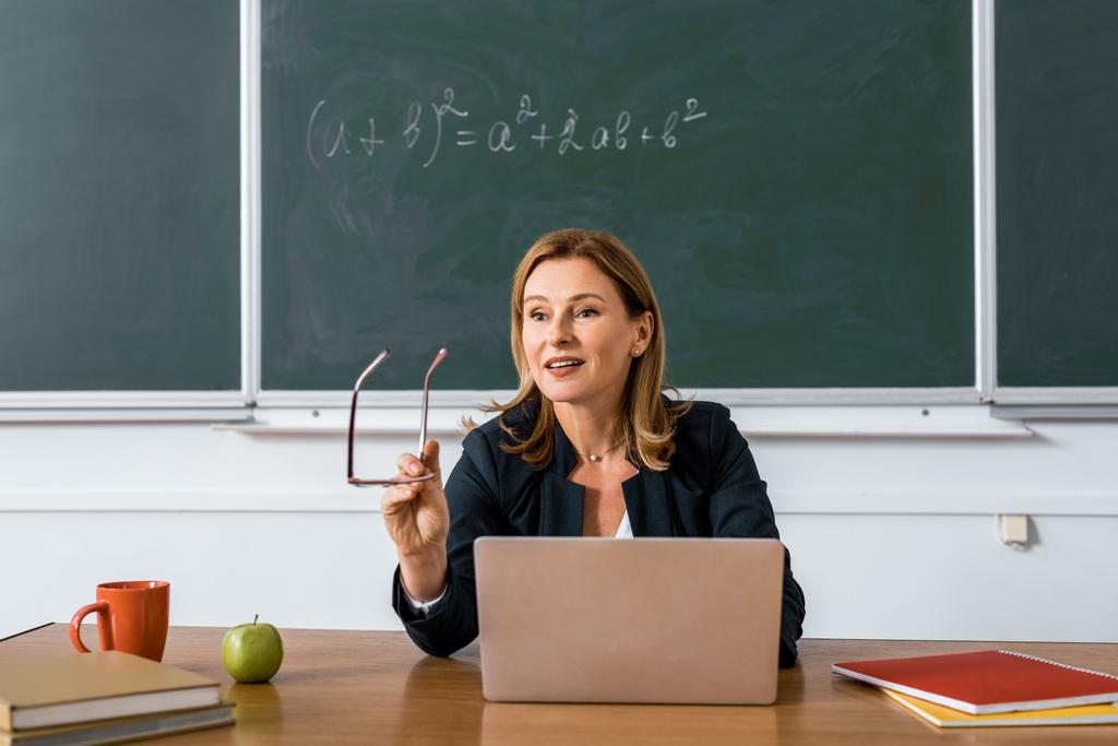δασκάλα, κάθεται στο γραφείο του υπολογιστή, κρατώντας γυαλιά και μιλώντας στην τάξη  - Φωτογραφία, εικόνα