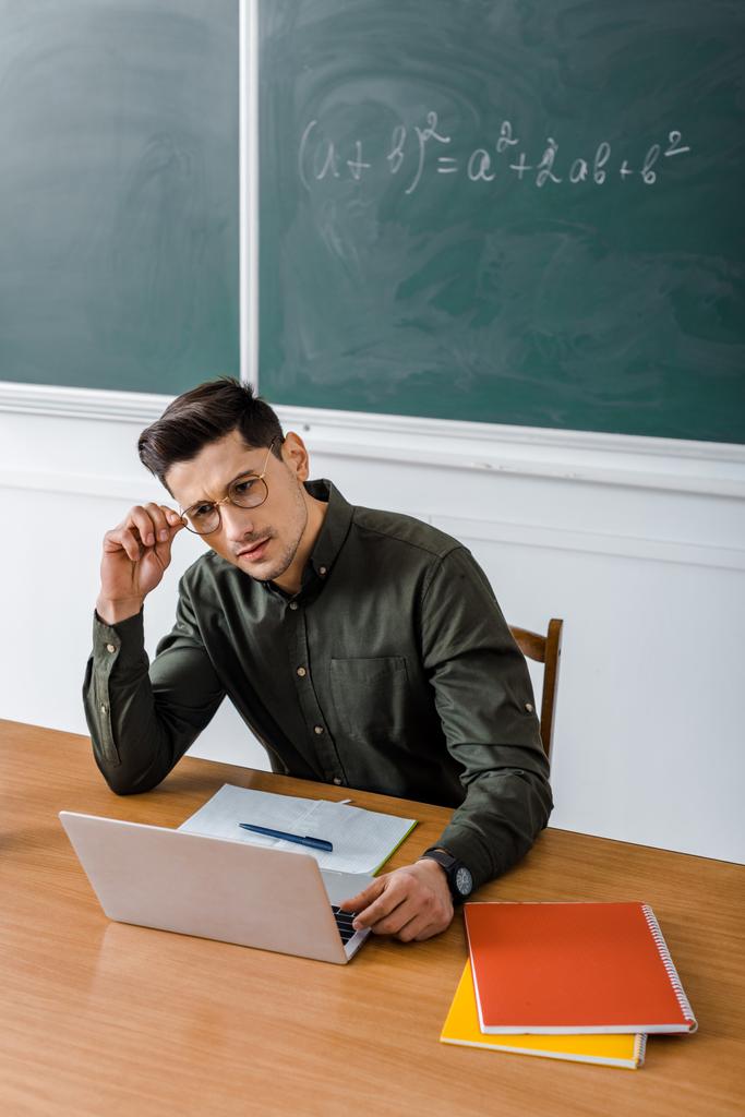 zamyšlený muž učitel v brýlích sedí a pomocí přenosného počítače na přepážce v učebně - Fotografie, Obrázek