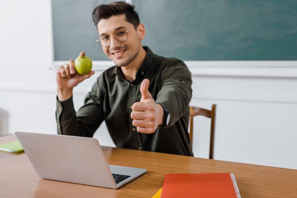 uśmiechnięty mężczyzna nauczyciel w okularach trzyma jabłko i pokazuje kciuk znak na biurko komputerowe w klasie - Zdjęcie, obraz