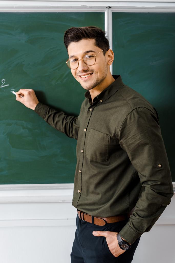 smiling male teacher looking at camera and writing on chalkboard in class - Photo, Image