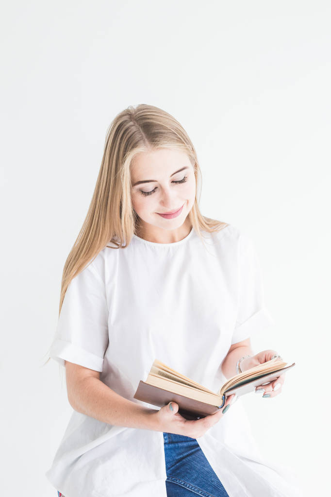 Portret van een jonge stijlvolle blond meisje in een witte T-shirt en spijkerbroek lezen van een boek op een witte achtergrond - Foto, afbeelding