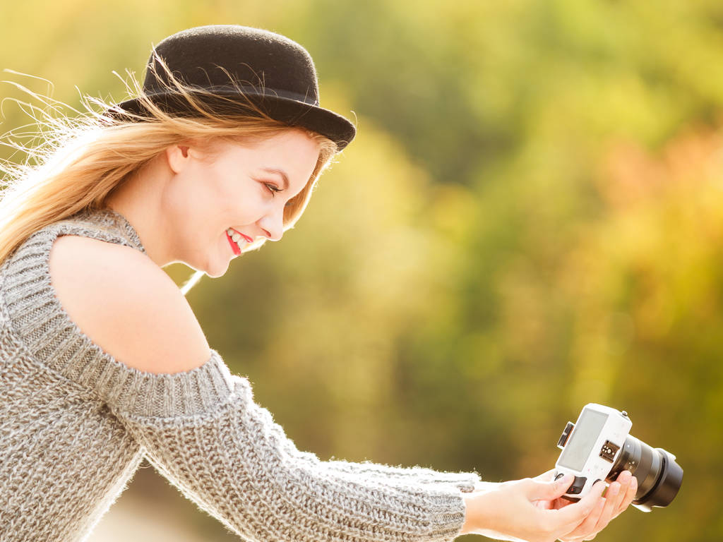 Улыбающаяся женщина в черной шляпе и сером свитере фотографирует с помощью цифровой камеры на открытой фотосессии. У женщины есть страсть
. - Фото, изображение