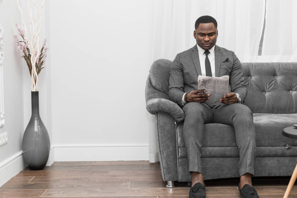 Молодой афроамериканский бизнесмен в сером костюме читает газету, сидя на диване
. - Фото, изображение