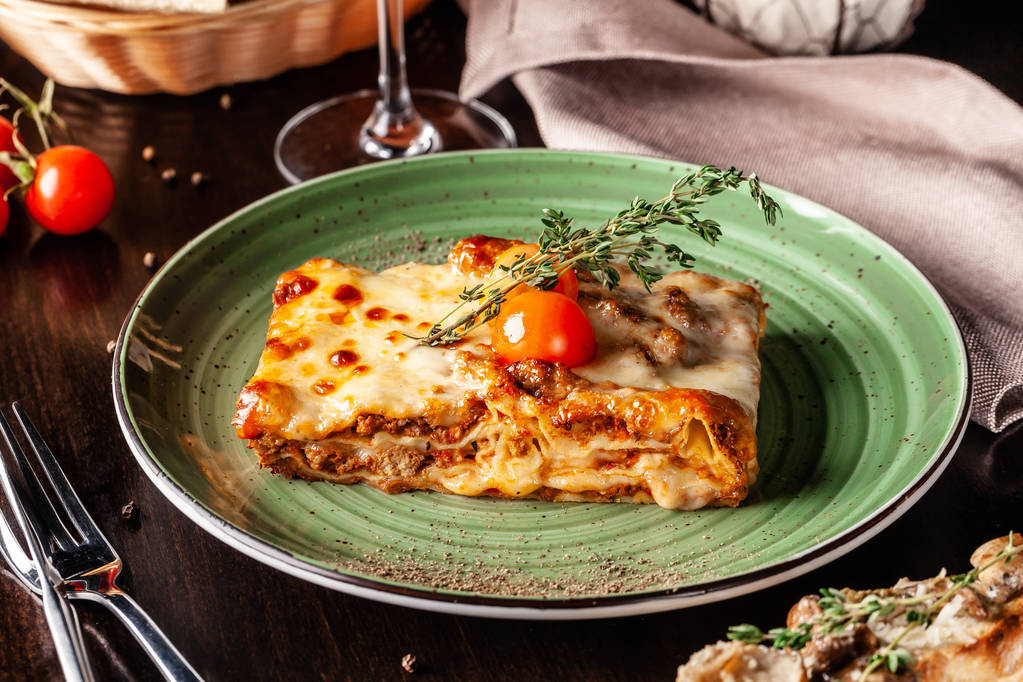 Koncepcja dania kuchni włoskiej. Pieczone lasagne z mielonego bolognese, makarony, pomidory cherry leży na zielony talerz w restauracji. Kieliszek czerwonego wina i chleba kosz na stole - Zdjęcie, obraz