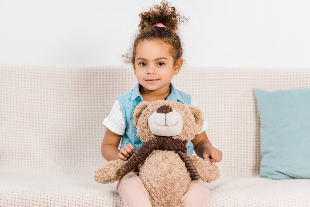 adorable african american child sitting on couch with teddy bear and smiling at camera - Photo, Image