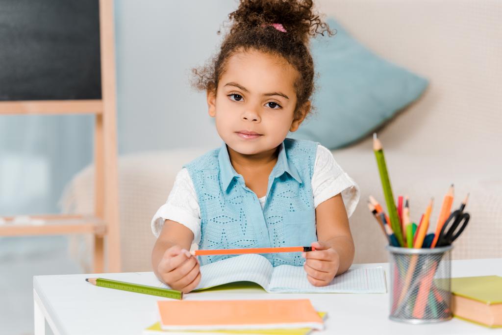 adorable enfant afro-américain tenant un crayon et regardant la caméra tout en écrivant dans le classeur
 - Photo, image