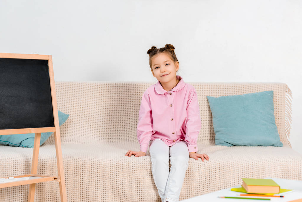 Вид в полный рост красивого маленького ребенка, сидящего на диване и улыбающегося перед камерой
  - Фото, изображение
