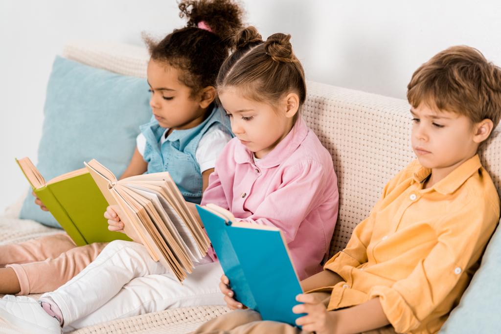 adorables niños multiétnicos sentados en un sofá y leyendo libros
  - Foto, Imagen
