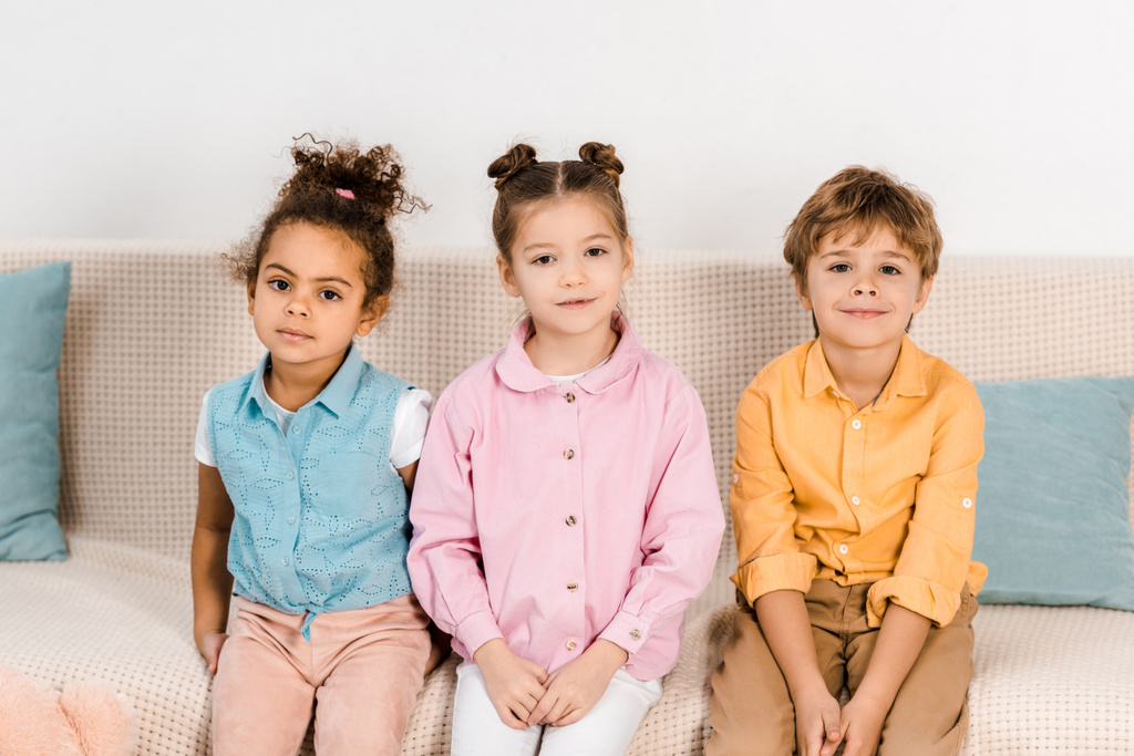 entzückende multiethnische Kinder, die auf dem Sofa sitzen und in die Kamera lächeln - Foto, Bild