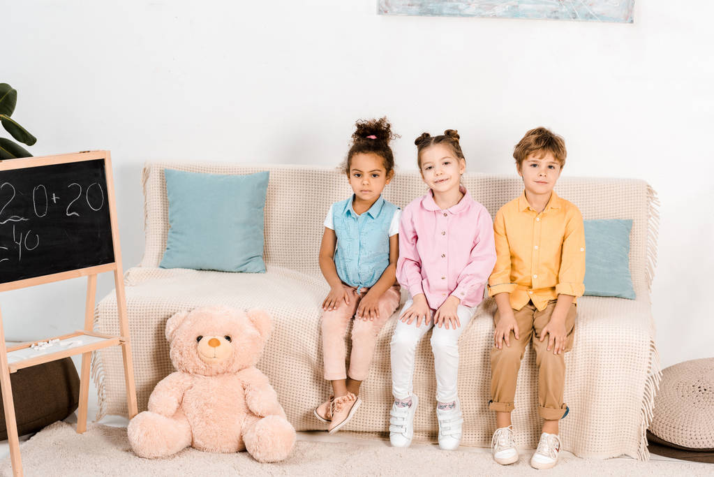 чарівні маленькі багатоетнічні діти сидять на дивані і дивляться на камеру
  - Фото, зображення