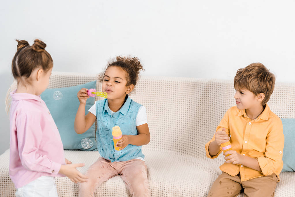 adorável multiétnico crianças sentado no sofá e soprando bolhas de sabão
  - Foto, Imagem