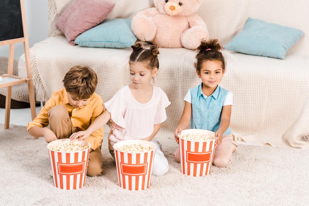 vysoký úhel pohled roztomilé mnohonárodnostní děti sedí na koberci a jíst popcorn z krabice - Fotografie, Obrázek
