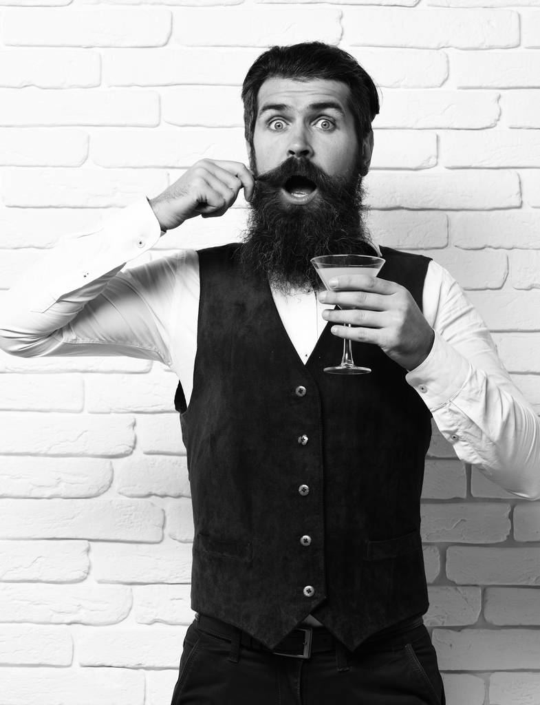 przystojny brodaty mężczyzna z długą brodą i wąsami ma stylowe włosy na zaskoczonej twarzy trzymając szklankę napoju alkoholowego w vintage zamszu skóra kamizelka o biały cegły ściany studio tło - Zdjęcie, obraz