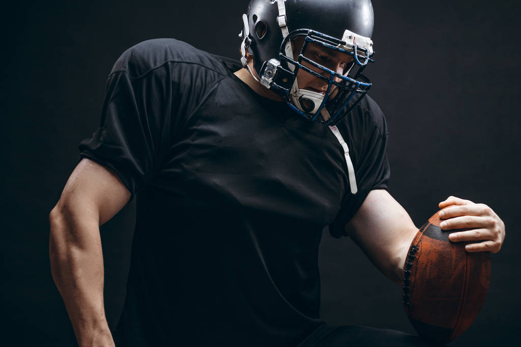 Αμερικανός ποδοσφαιριστής σε μαύρο αθλητικό ένδυμα με μπάλα σε μαύρο φόντο - Φωτογραφία, εικόνα