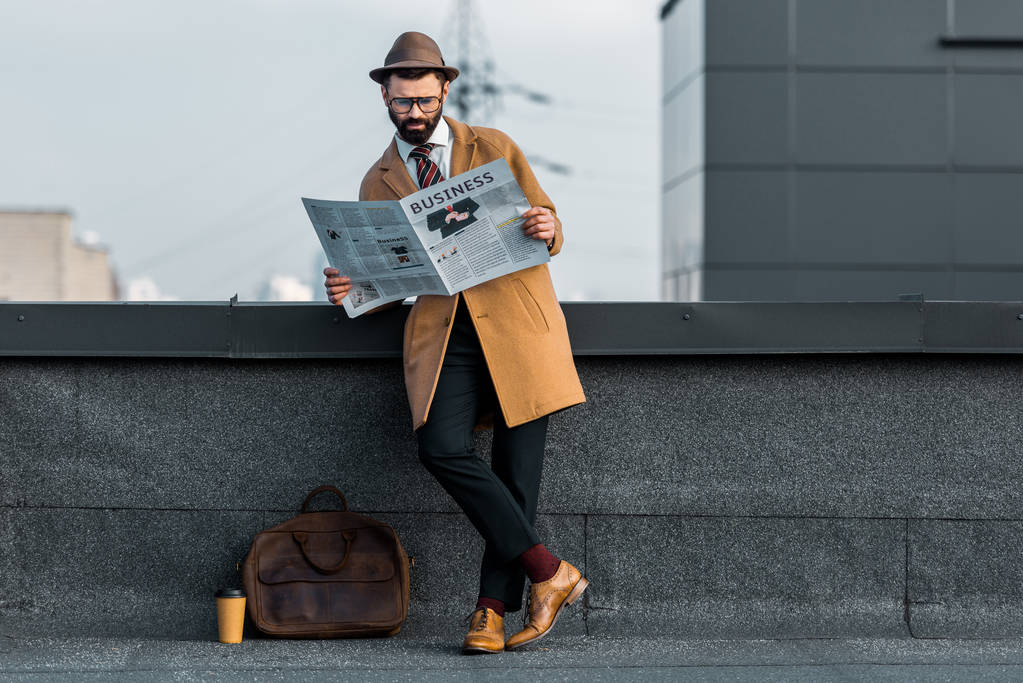 組んだ足の側に立って、屋根の上のビジネス新聞を読む大人の髭の男 - 写真・画像