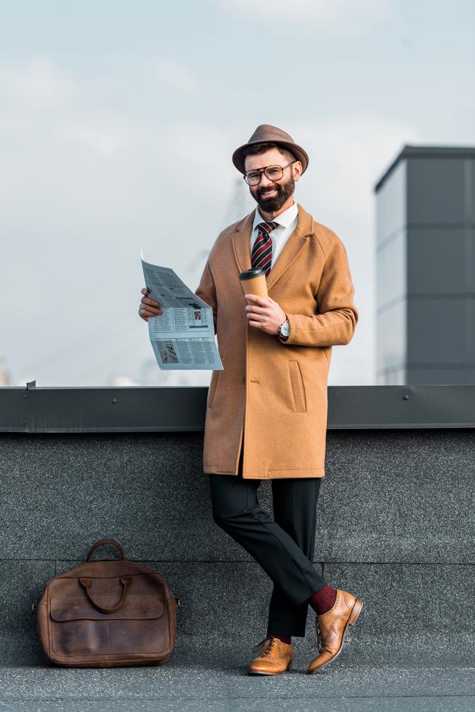 χαμογελώντας γενειοφόρος επιχειρηματίας που στέκεται με σταυρωμένα πόδια κρατώντας την εφημερίδα και καφέ για να πάει - Φωτογραφία, εικόνα