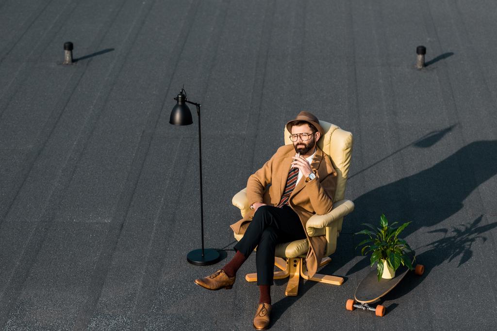 uomo d'affari adulto che riposa in poltrona con sigaretta vicino alla lampada e pianta su penny board
 - Foto, immagini
