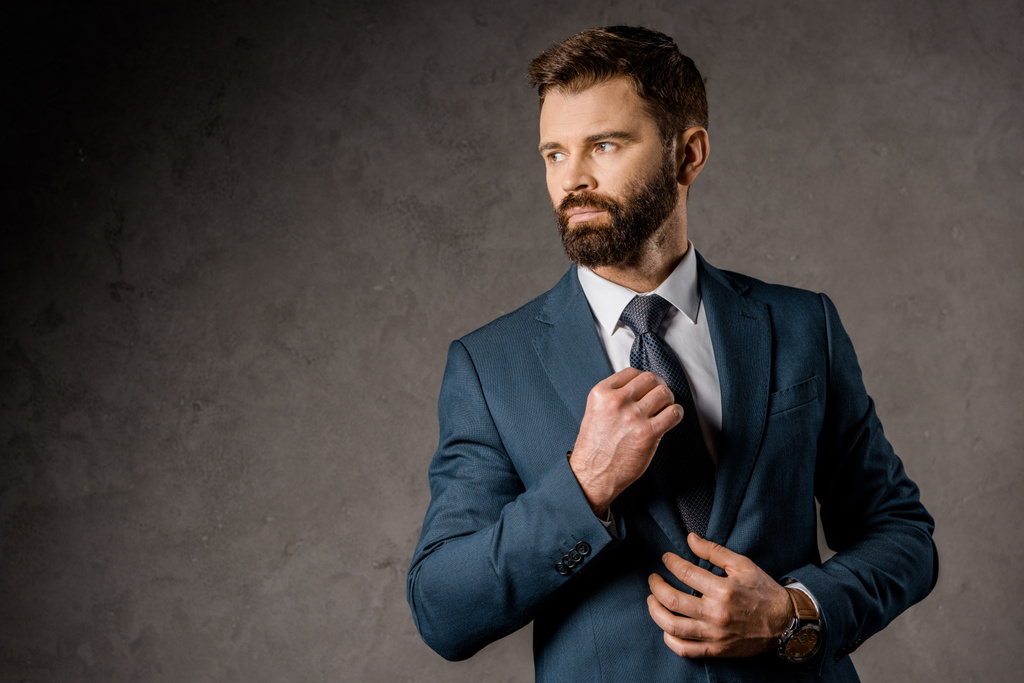confident bearded businessman standing in formalwear and touching tie  - Photo, Image