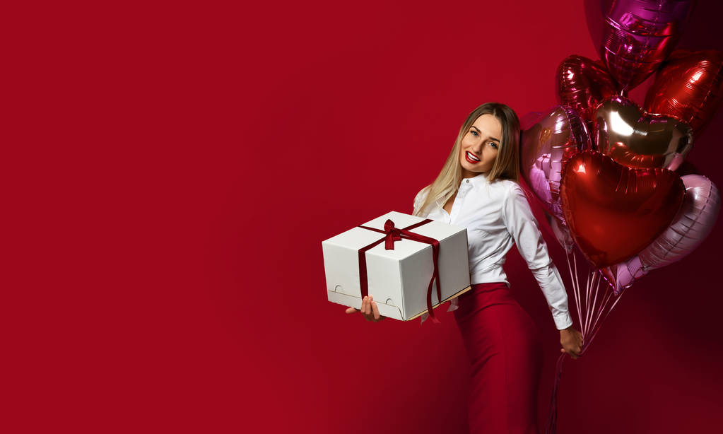 Женщина с подарочной коробкой и фиолетовым розовым сердечком счастливо улыбается на красном фоне
 - Фото, изображение