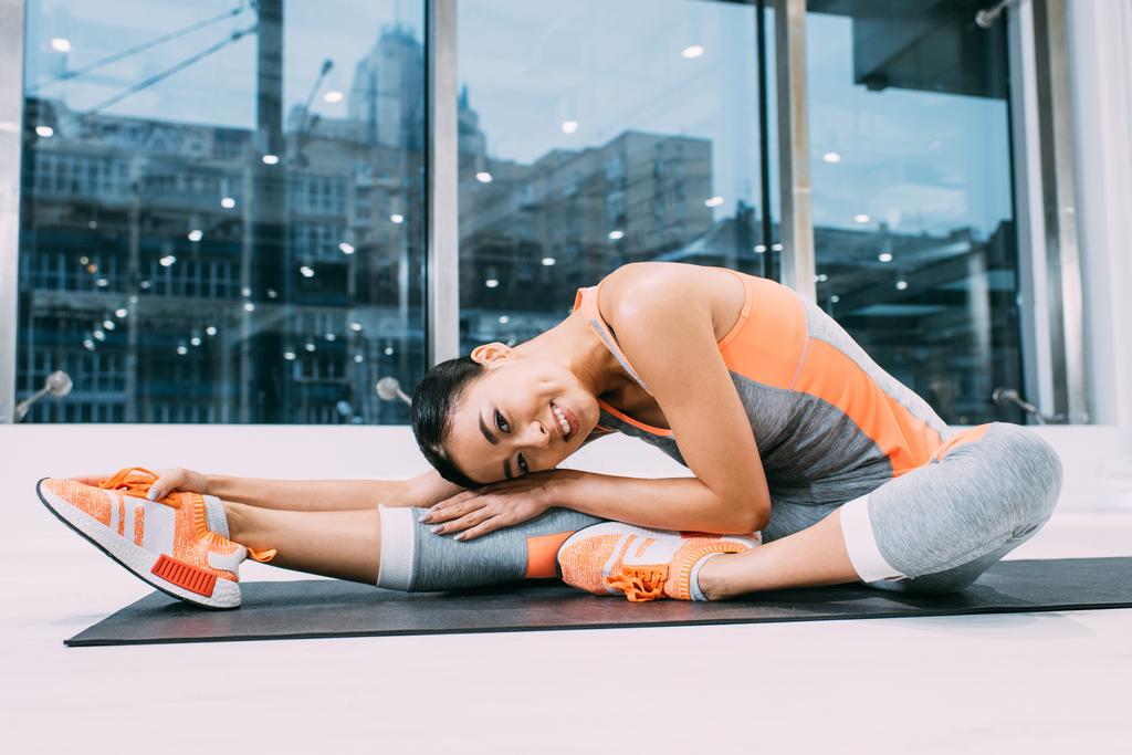 lächelnde asiatische Mädchen beim Stretching auf der Fitnessmatte im Fitnessstudio - Foto, Bild