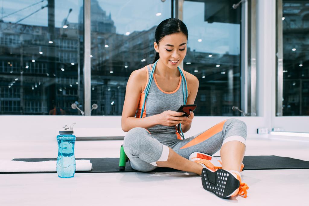 attractive asian girl sitting on fitness mat and using smartphone in sports center - Photo, Image