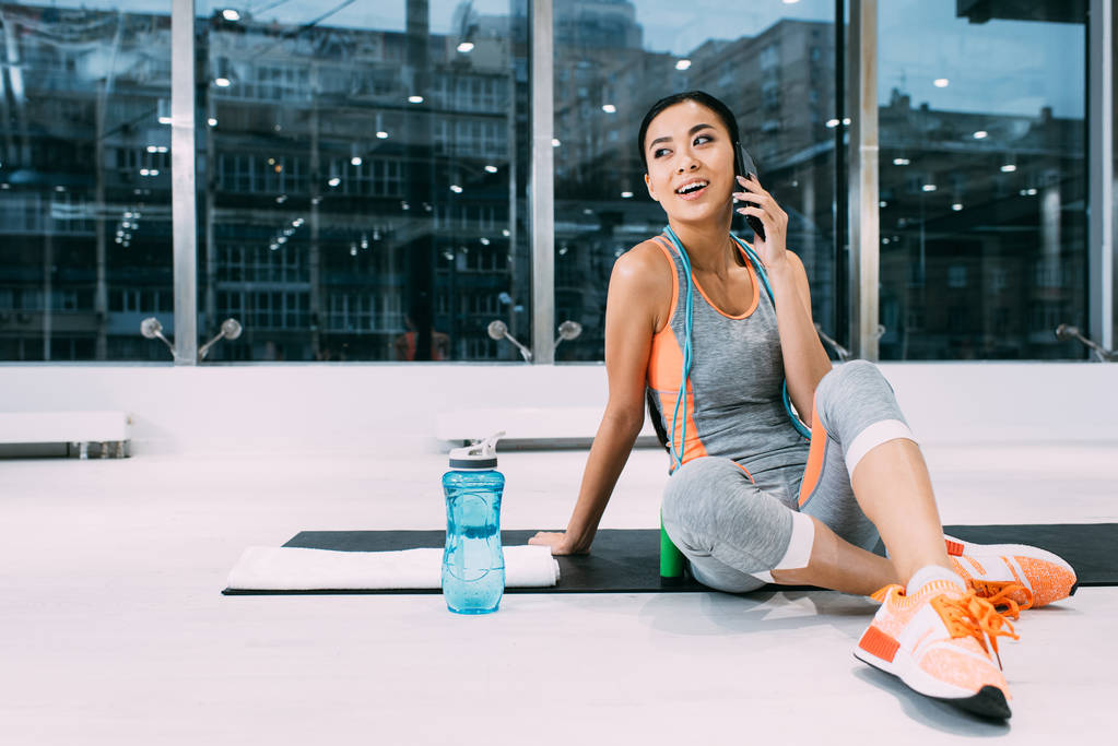 atrakcyjny azjatycki sportsmenka siedzi na fitness Mata i mówić na smartfonie w centrum sportowe - Zdjęcie, obraz