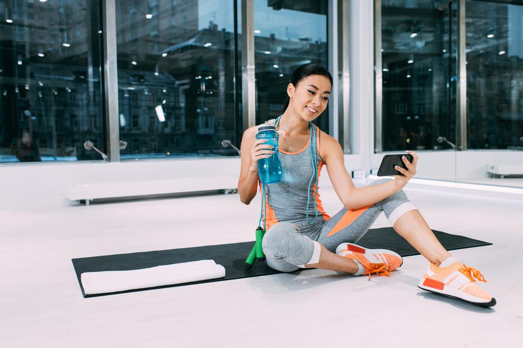 usmívající se Asijská sportovkyně sedí na fitness mat, držící sportovní láhev s vodou a pořizování selfie v tělocvičně - Fotografie, Obrázek