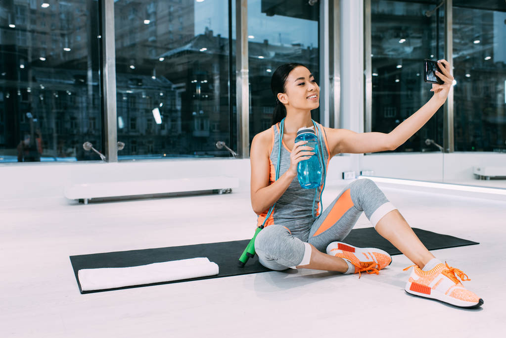 усміхнена азіатська дівчина сидить на фітнес-маті, тримає спортивну пляшку з водою і приймає селфі в спортзалі
 - Фото, зображення