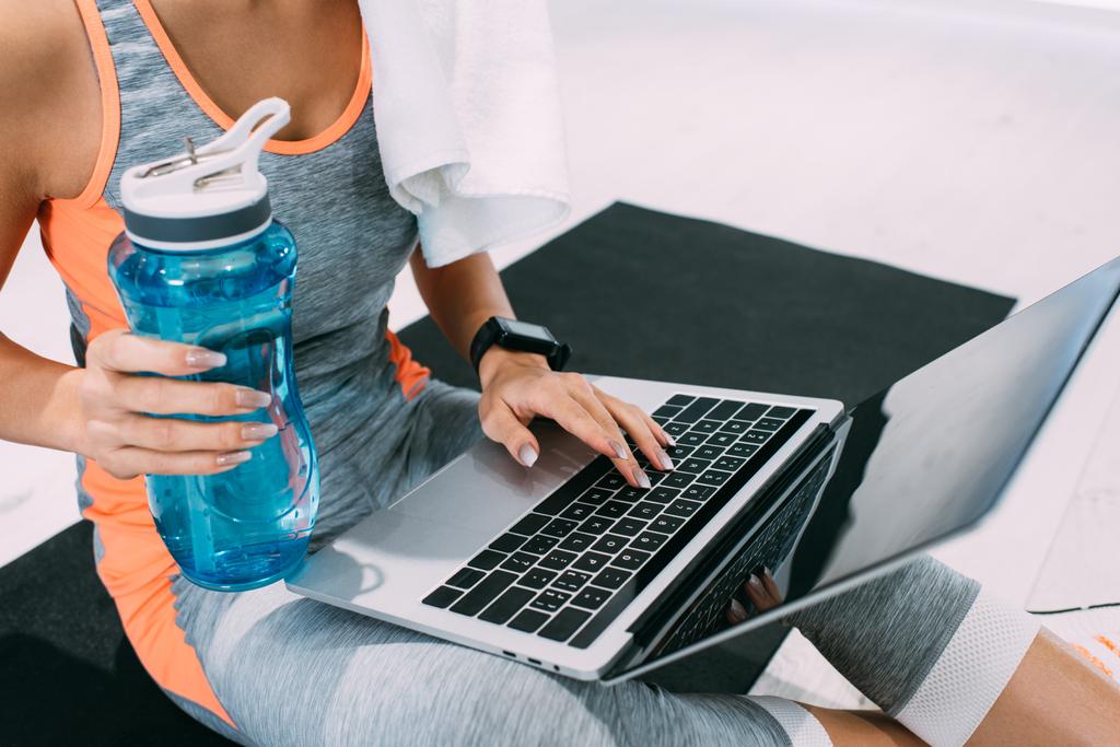 vue recadrée d'une fille assise sur un tapis de fitness, tapant sur un clavier d'ordinateur portable et tenant une bouteille de sport au gymnase
 - Photo, image