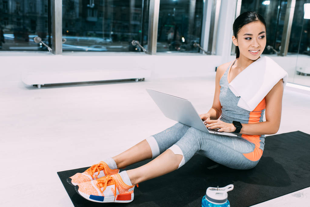 mladá Asijská sportovkyně s ručníkem na ramenou sedí na fitness mat, psaní na klávesnici notebooku a usmívající se v tělocvičně - Fotografie, Obrázek