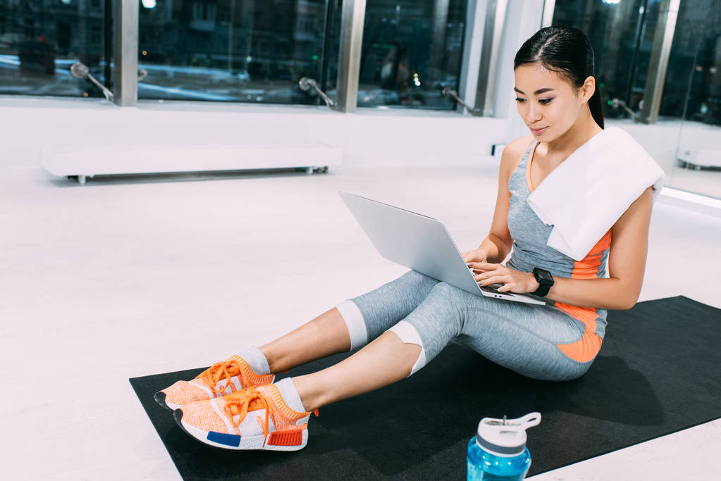 Atrakcyjna dziewczyna azjatyckich z ręcznikiem na barki siedząc na fitness Mata i wpisując na klawiaturze laptopa siłowni - Zdjęcie, obraz