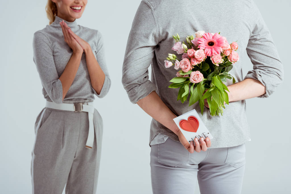 vista ritagliata della donna con le mani piegate guardando l'uomo nascondendo bouquet di fiori e biglietto di auguri di San Valentino dietro la schiena isolato sul grigio
 - Foto, immagini