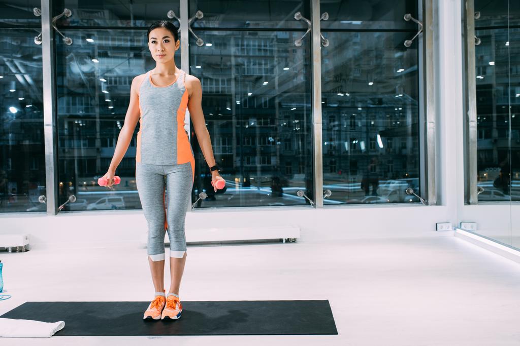 привлекательная азиатская спортсменка, стоящая на фитнес-коврике и делающая упражнения с гантелями в тренажерном зале
 - Фото, изображение