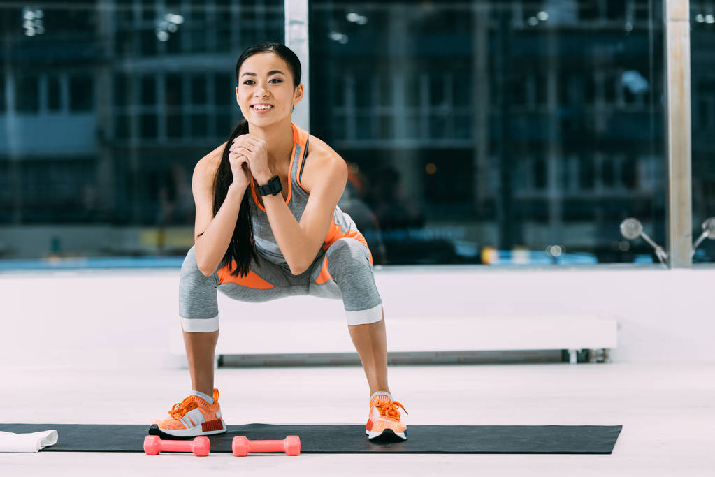 sonriente asiático chica haciendo sentadillas en fitness mat en gimnasio
 - Foto, Imagen