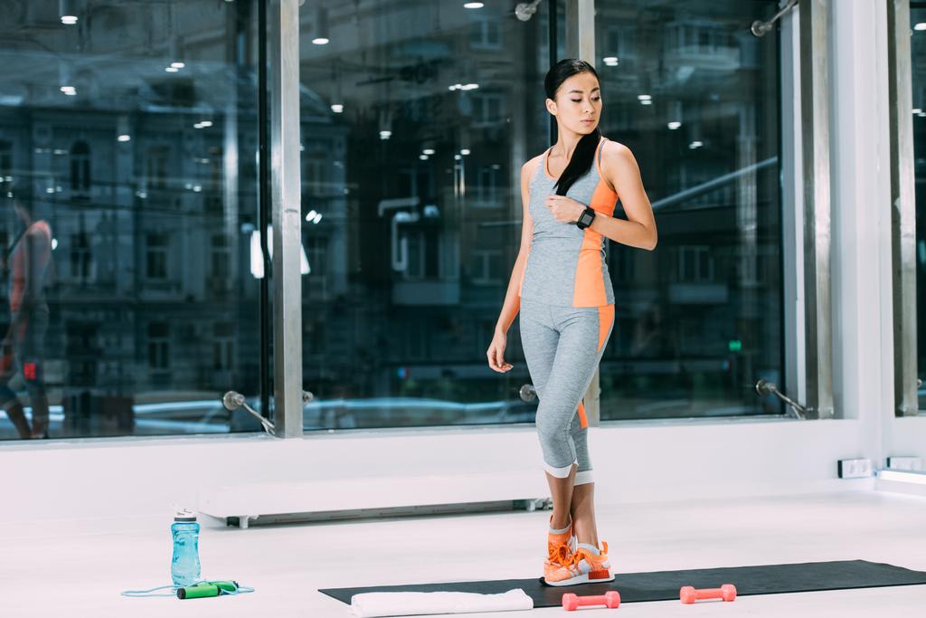 slim asian girl standing on fitness mat and touching hair in sports center - Photo, Image