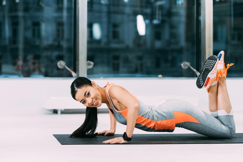 attraktive asiatische Sportlerin macht Liegestütze auf Fitnessmatte und lächelt im Fitnessstudio - Foto, Bild