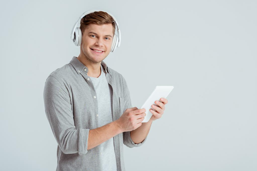 homem em fones de ouvido ouvindo música, olhando para a câmera e usando tablet digital isolado em cinza
 - Foto, Imagem