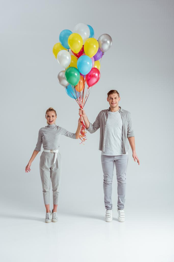 usmívající se pár stojící na špičkách, při pohledu na fotoaparát a drží svazek barevných balónků na šedém pozadí - Fotografie, Obrázek