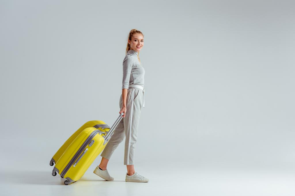 woman carrying yellow suitcases and looking at camera on grey background, travel concept - Photo, Image