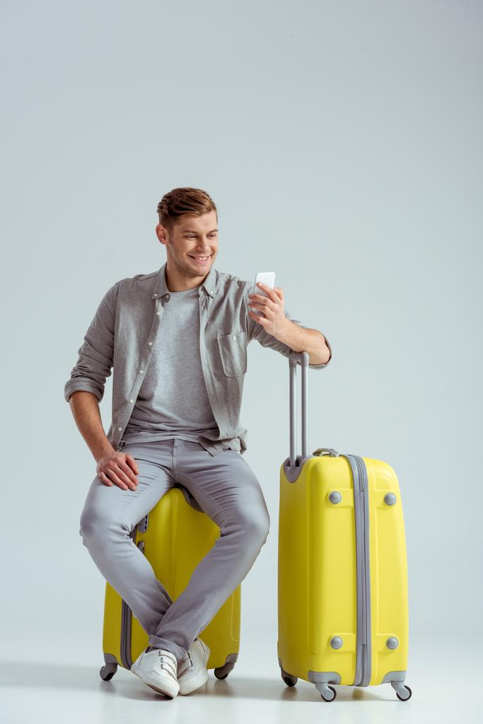 smiling man sitting on yellow suitcase and using smartphone on grey background, travel concept - Photo, Image