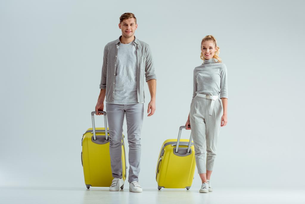 улыбающаяся пара с желтыми чемоданами и смотрящая на камеру на сером фоне, концепция путешествия
 - Фото, изображение