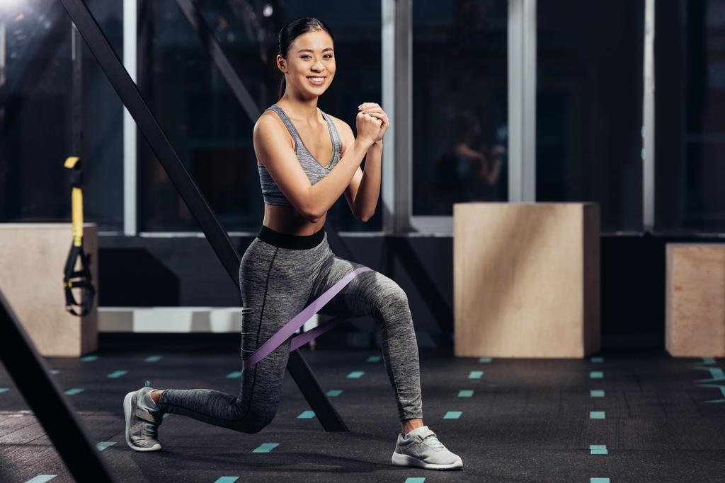 lächelnde asiatische Mädchen beim Ausfallschritt mit Gummibändern im Fitnessstudio  - Foto, Bild