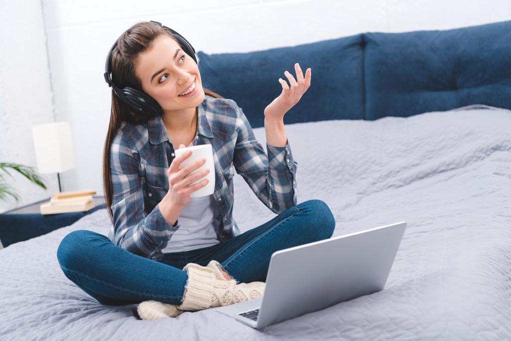 beautiful happy girl in headphones holding cup and using laptop on bed    - Photo, Image