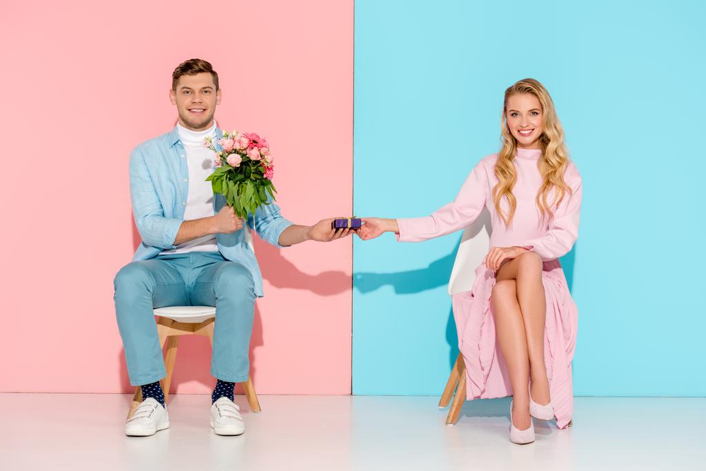 пара сидить на стільцях, коли чоловік представляє подарункову коробку з квітковим букетом усміхненій жінці на рожевому та синьому тлі
 - Фото, зображення