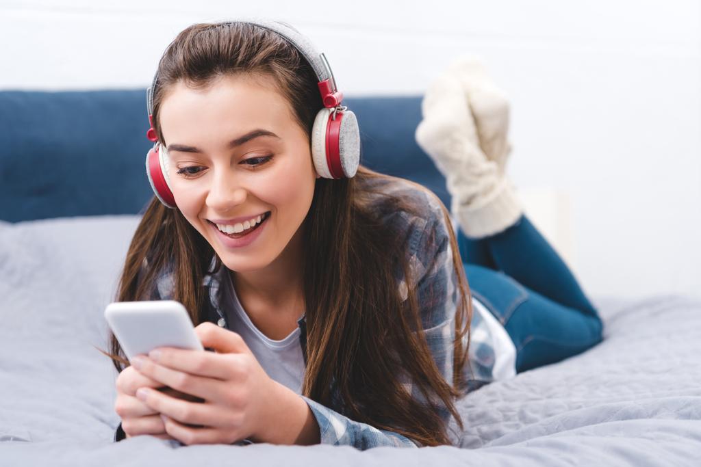 beautiful smiling young woman in headphones lying on bed and using smartphone - Photo, Image