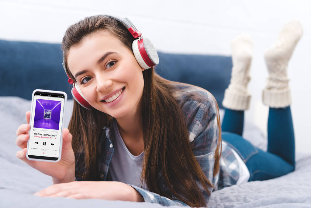 приваблива дівчина в навушниках проведення смартфон з програми музика і посміхатися камери лежачи на ліжку   - Фото, зображення