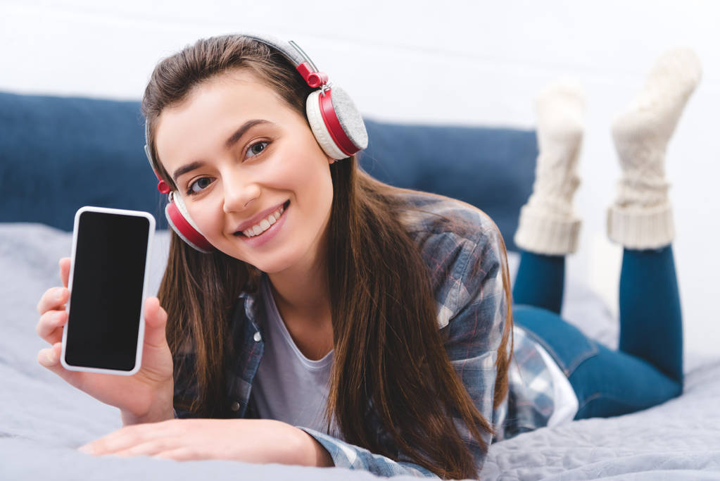 piękna młoda kobieta w słuchawkach, trzymając smartfon z pustego ekranu i uśmiecha się do kamery leżąc na łóżku  - Zdjęcie, obraz