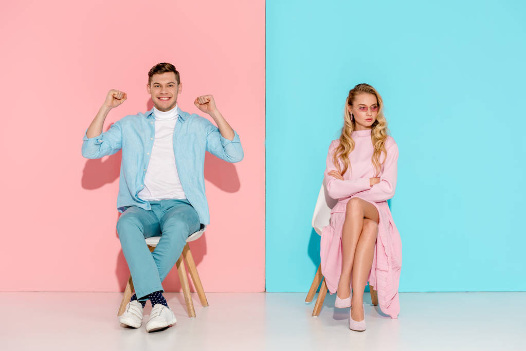 Unzufriedene Frau mit verschränkten Armen und jubelnder Mann mit geballten Fäusten, die auf Stühlen auf rosa und blauem Hintergrund sitzen - Foto, Bild