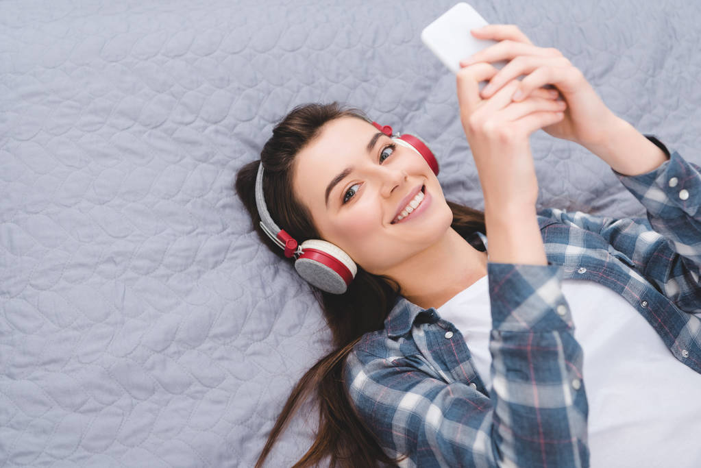 glückliche junge Frau mit Kopfhörer, Smartphone im Bett und lächelnd in die Kamera - Foto, Bild