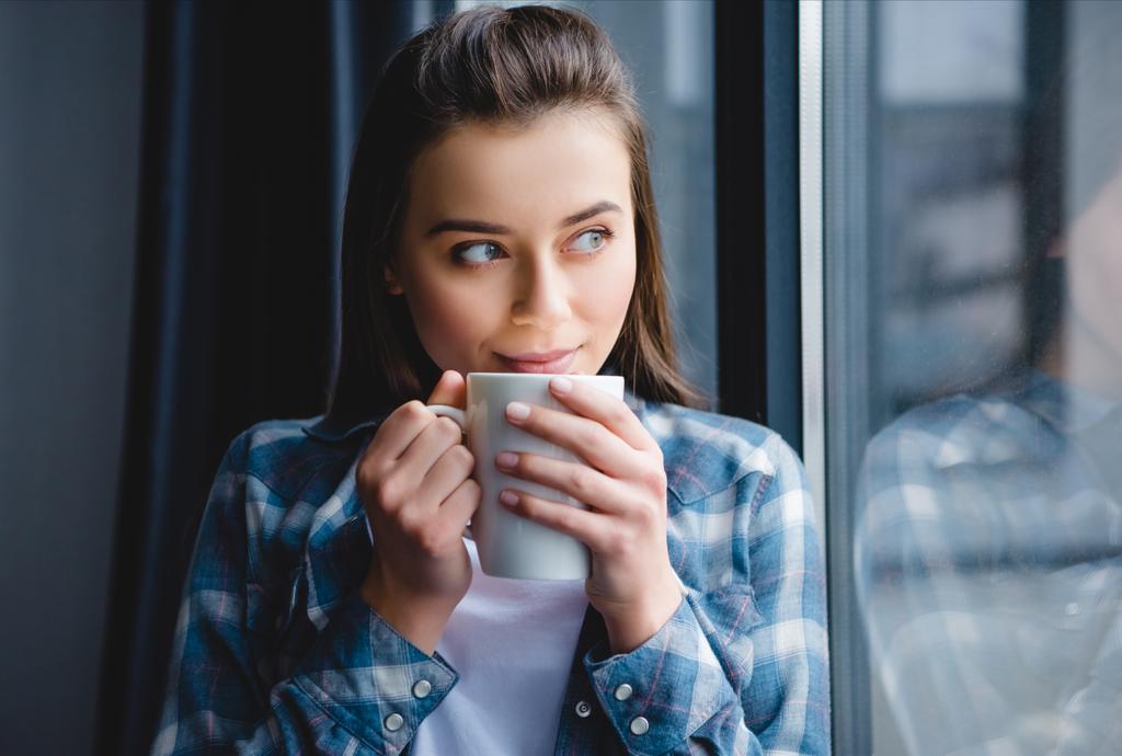 όμορφη νεαρή ευτυχισμένη γυναίκα πίνοντας τσάι και εξετάζοντας το παράθυρο  - Φωτογραφία, εικόνα